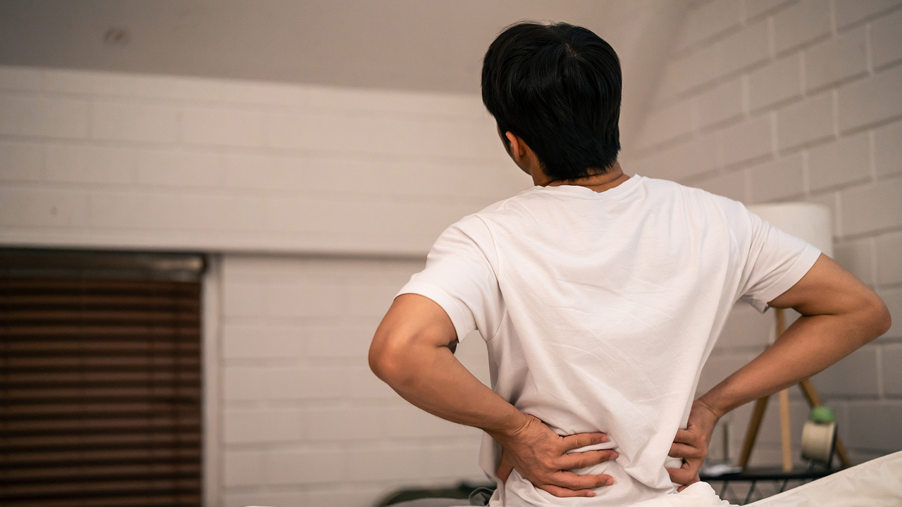 Man experiencing lower back pain while sitting on a bed.