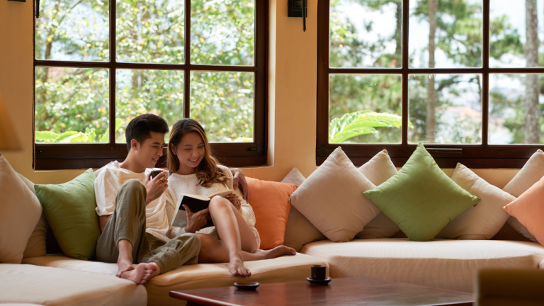Young couple relaxing together on a cozy sofa, reading a book and enjoying a peaceful moment in a bright living room overlooking tropical greenery.