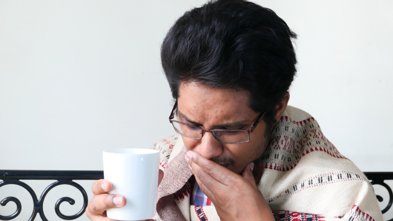 Man wearing glasses and wrapped in a blanket, covering his mouth while holding a white cup and feeling unwell.