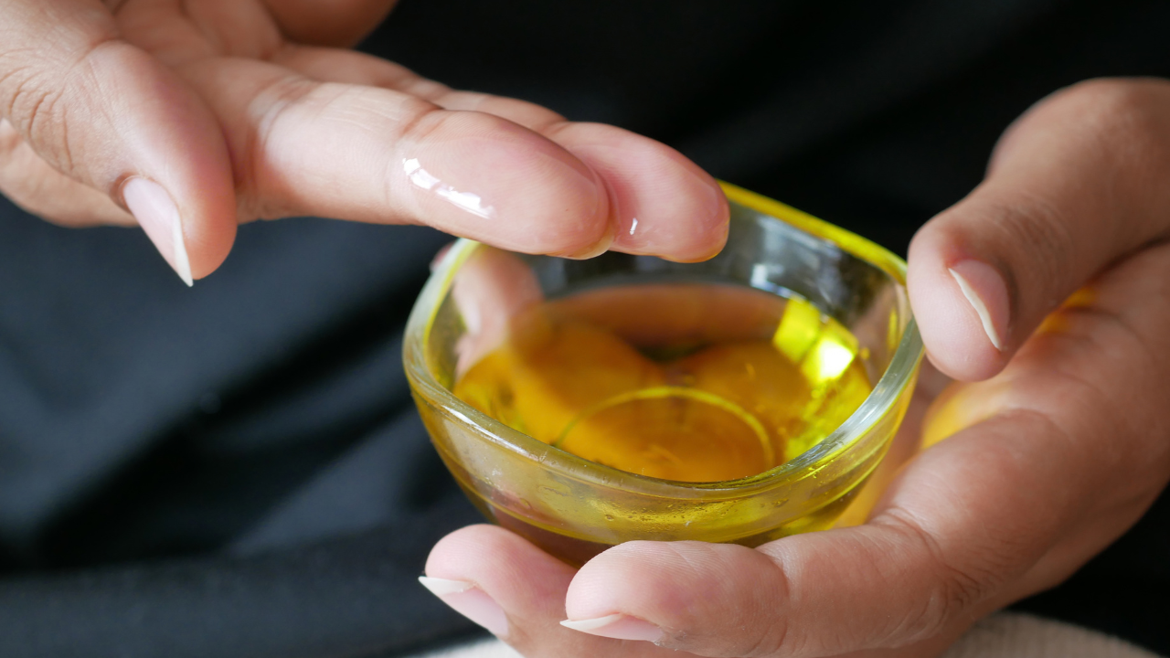 Close-up of hands holding a small glass bowl filled with golden herbal oil, with a drop of oil on a fingertip.